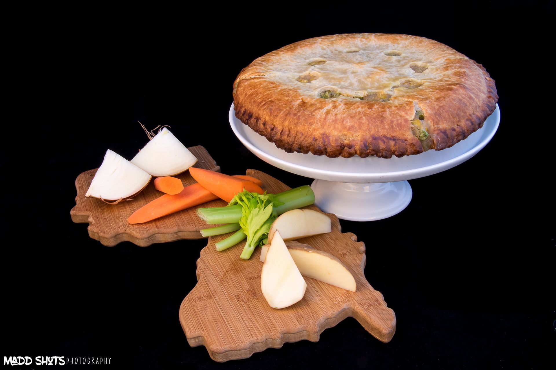 Chicken pot pie on a cake stand with assorted vegetables on wooden boards, black background.
