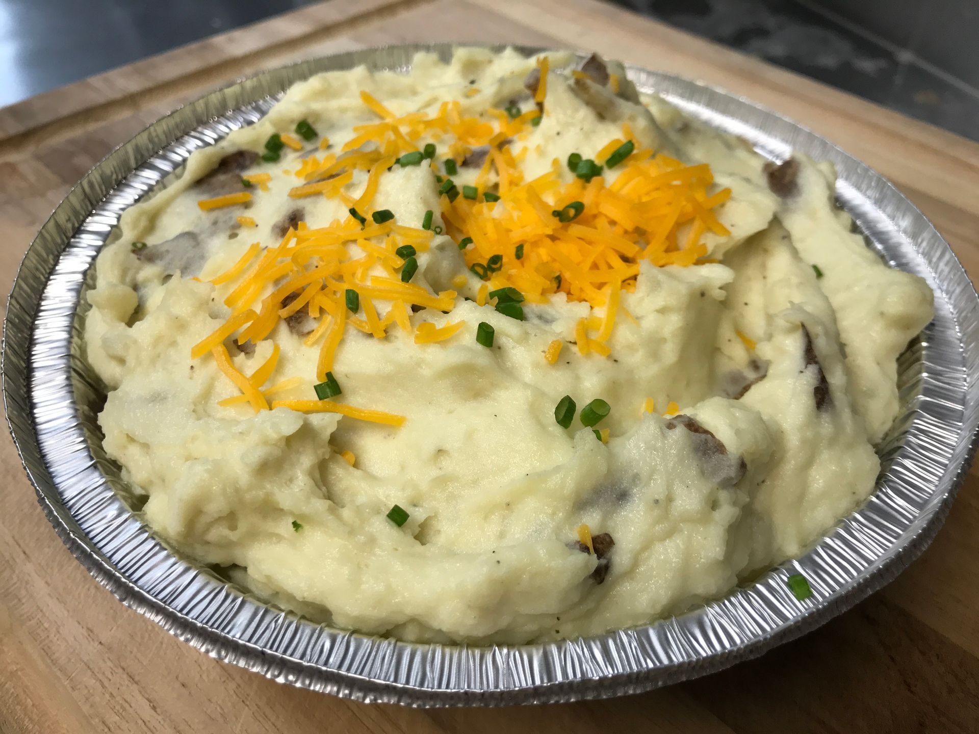 Mashed potatoes in a foil pie dish, topped with shredded cheddar cheese and chives.