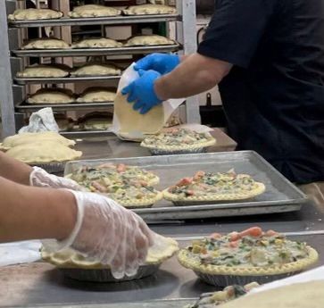 People preparing savory pies in a bakery. Filling being added and crusts are being placed. Racks of uncooked pies in the background.