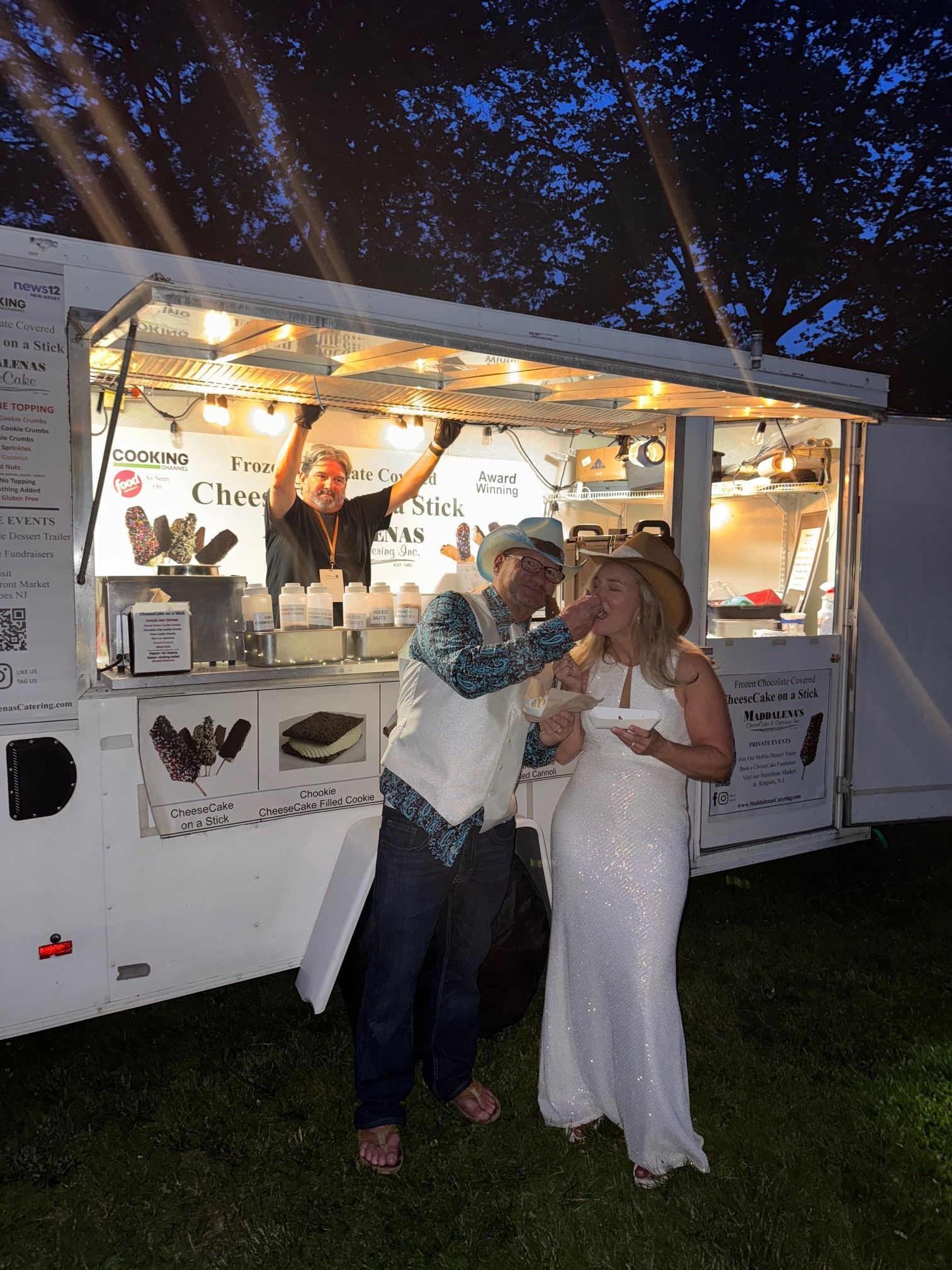 Couple in wedding attire feeding each other dessert from an ice cream truck at night.