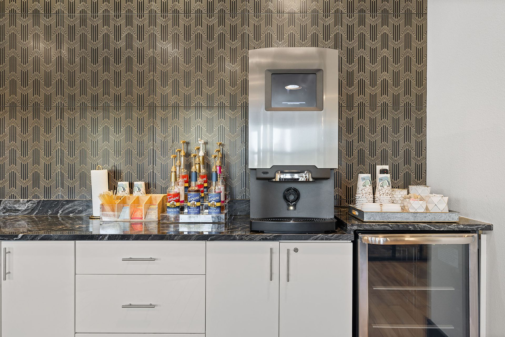 Hotel beverage station with coffee machine, drinks, cups, and snacks on a counter at Two99 Monroe.