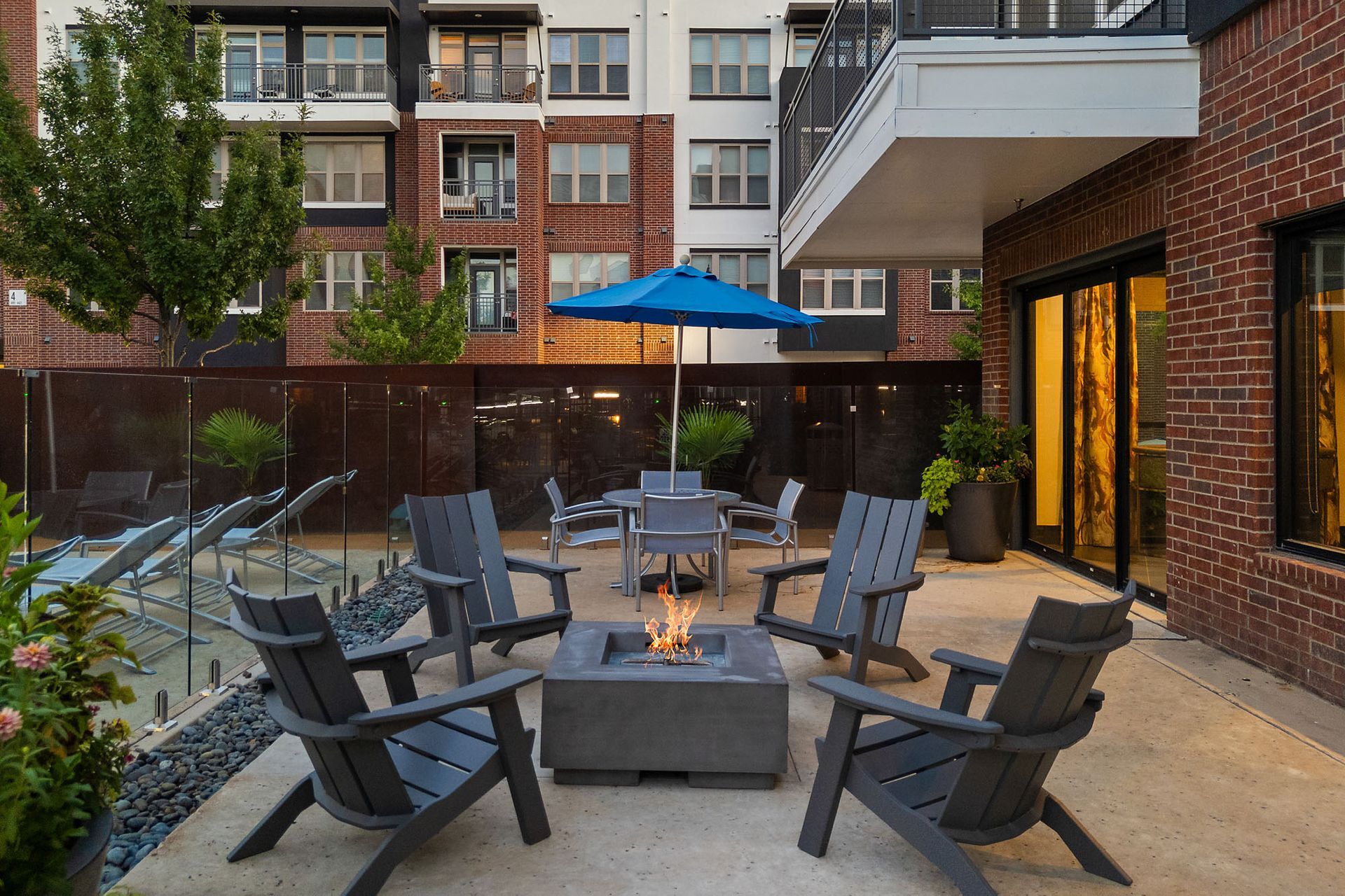 Outdoor patio with gray adirondack chairs, fire pit, and blue umbrellas beside apartment buildings at Two99 Monroe.