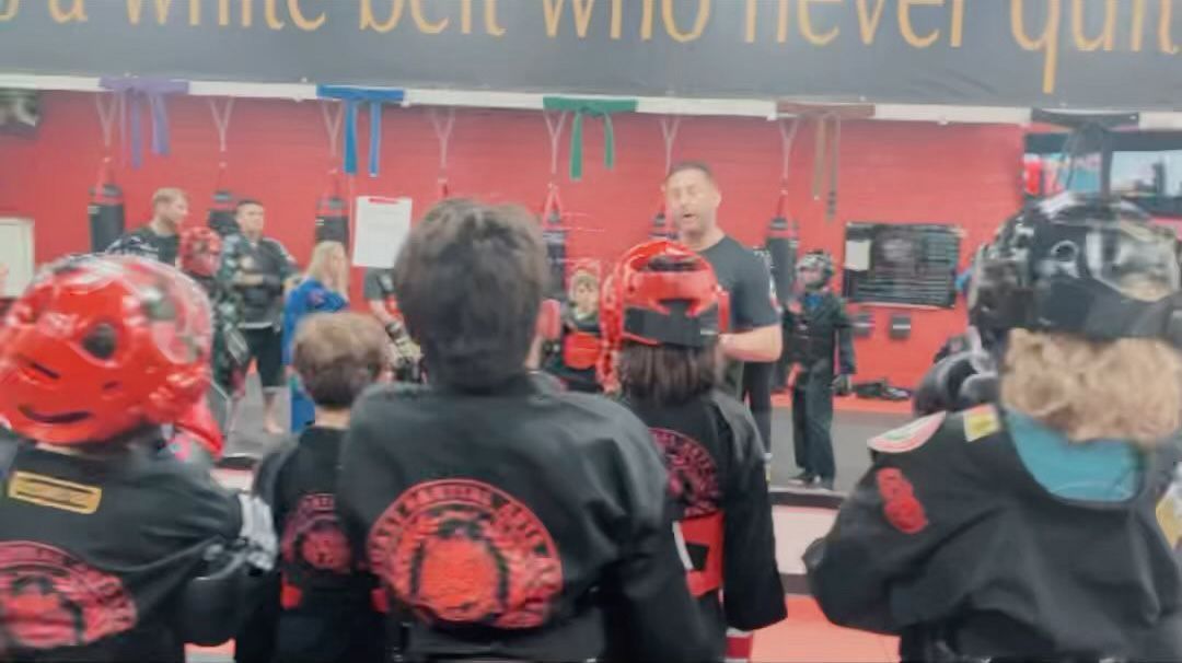 A group of children are standing in front of a man in a gym.