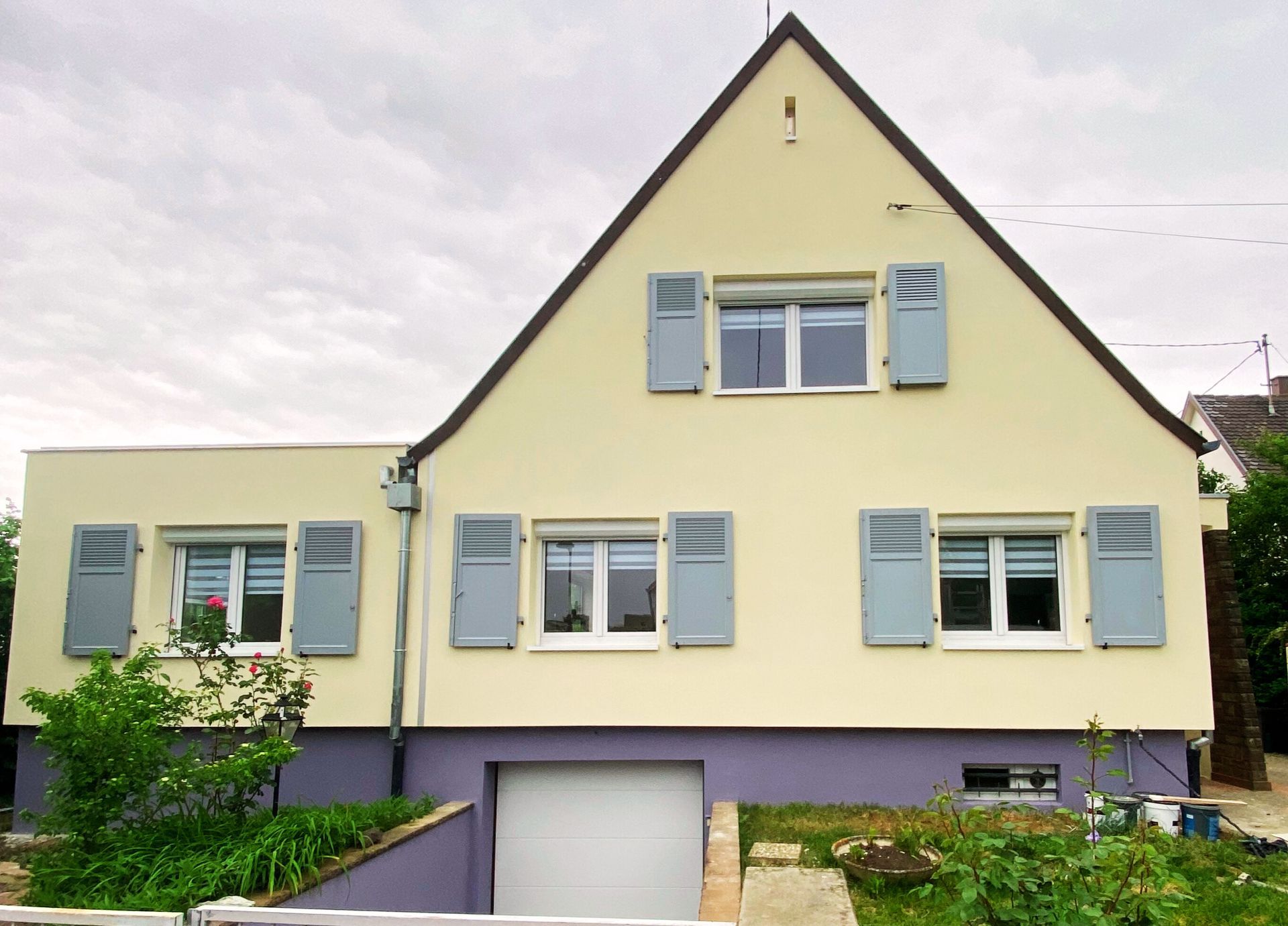 Maison jaune pastel avec volets bleus et garage, vue de face sous un ciel nuageux
