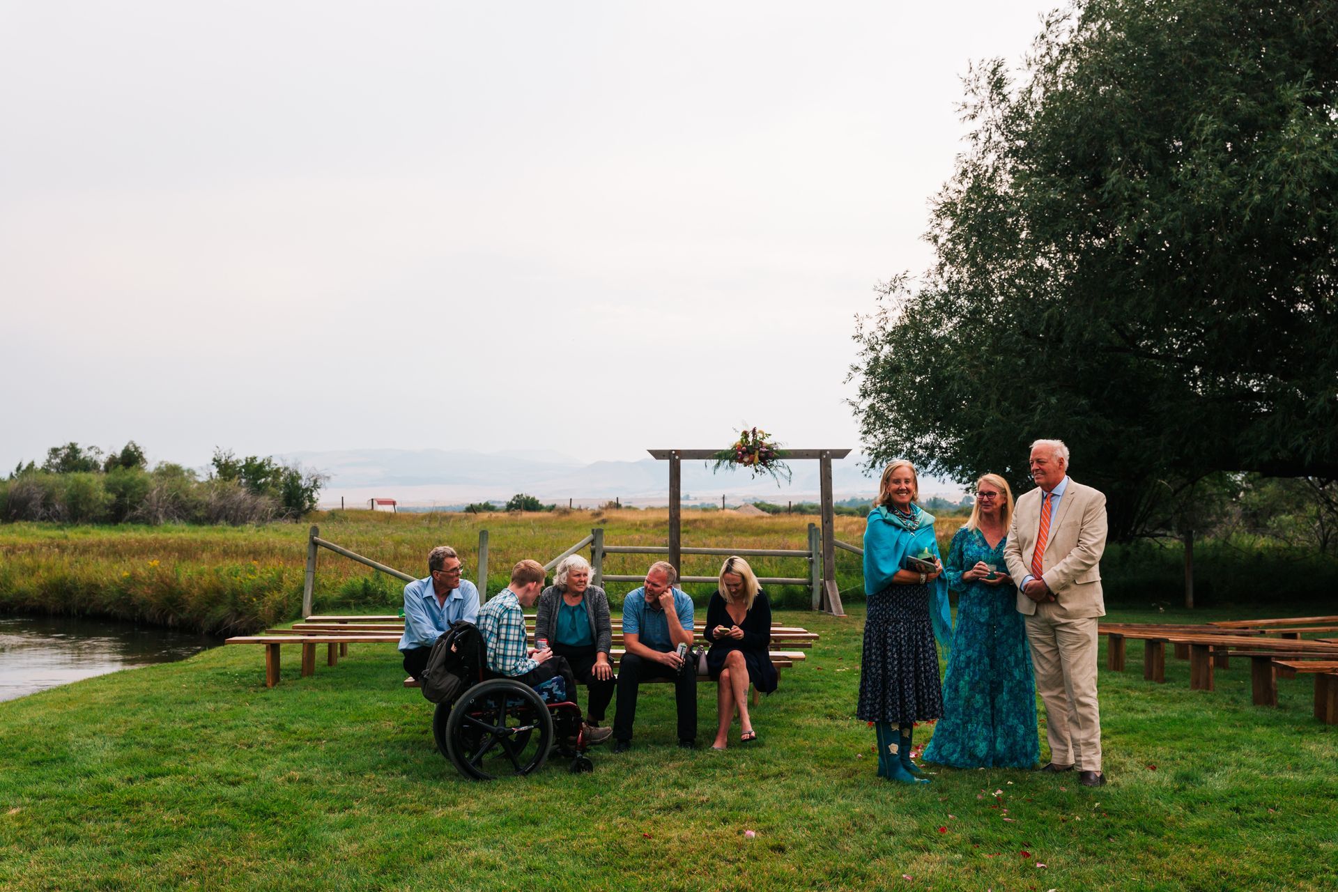 Group of people standing and sitting on a grassy lawn near a pond, with trees and a cloudy sky.