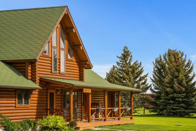 Wooden log cabin with green roof and porch on a sunny lawn, framed by tall trees.