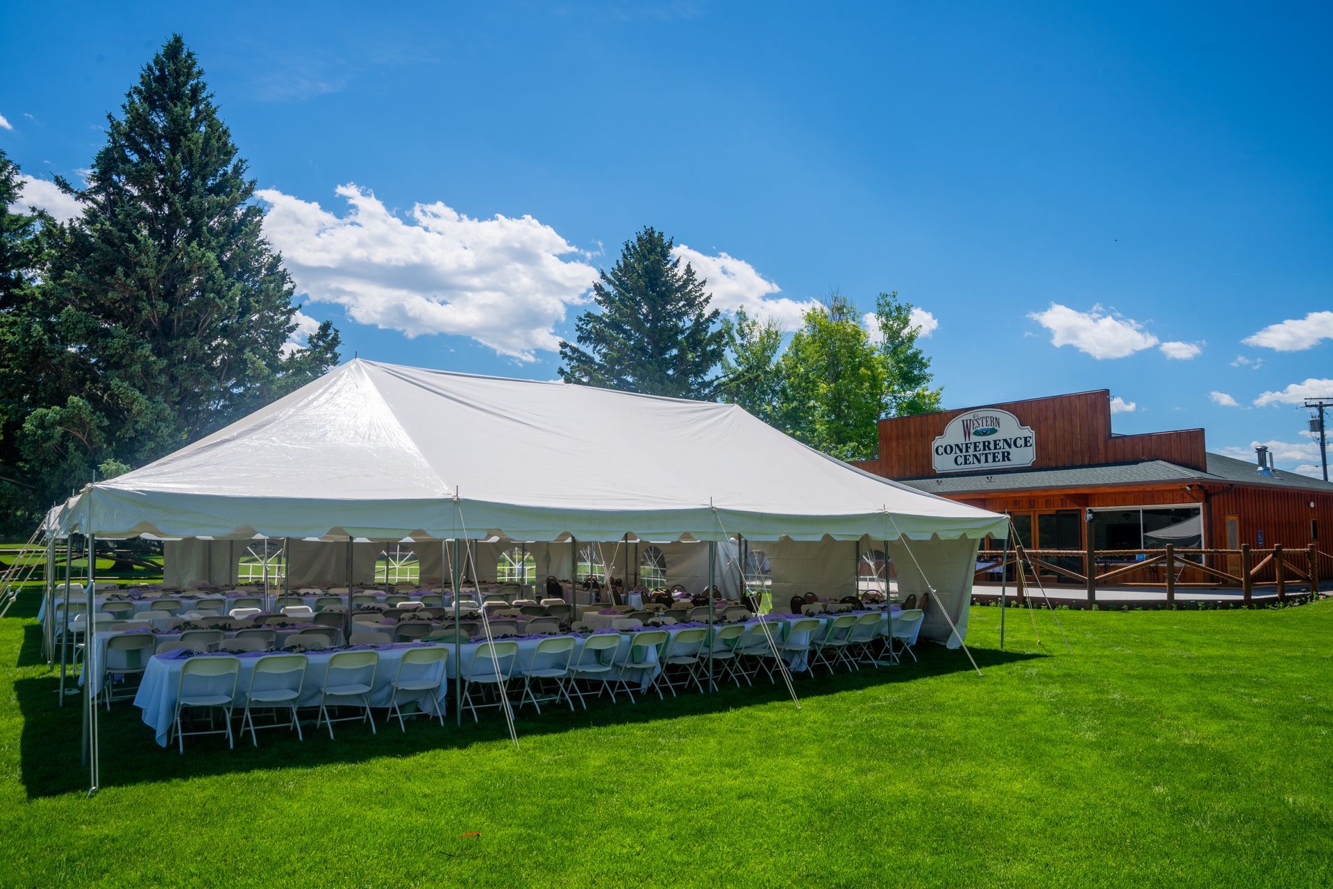 White event tent with seated guests on a green lawn near a brick building under a blue sky