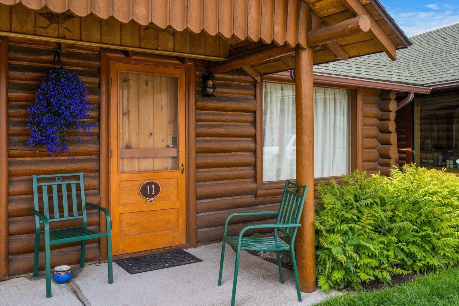 Wooden cabin porch with two green chairs, a wooden door, and a hanging basket of blue flowers.
