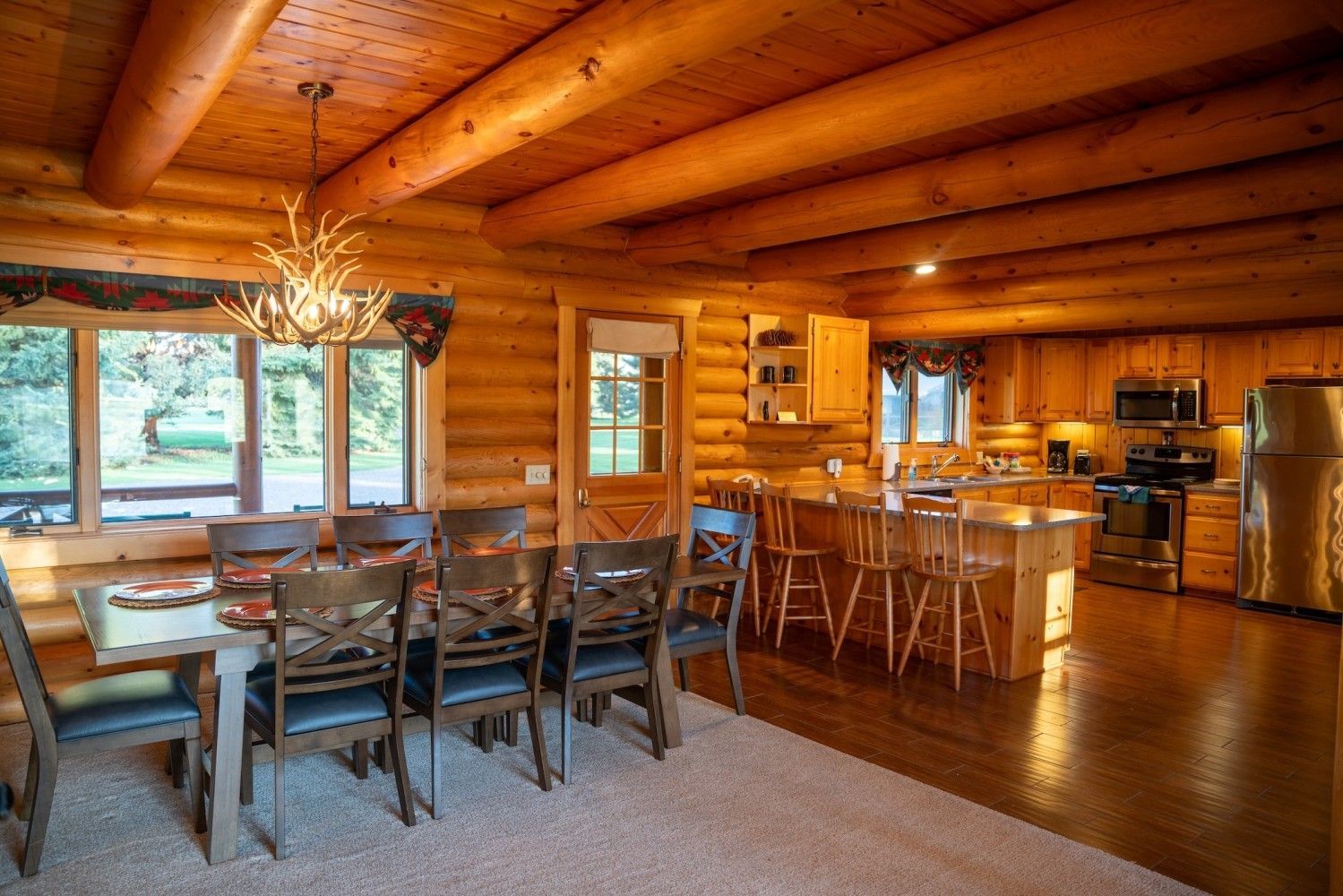 Rustic log cabin dining room and kitchen with wood-paneled walls, long table, and large windows
