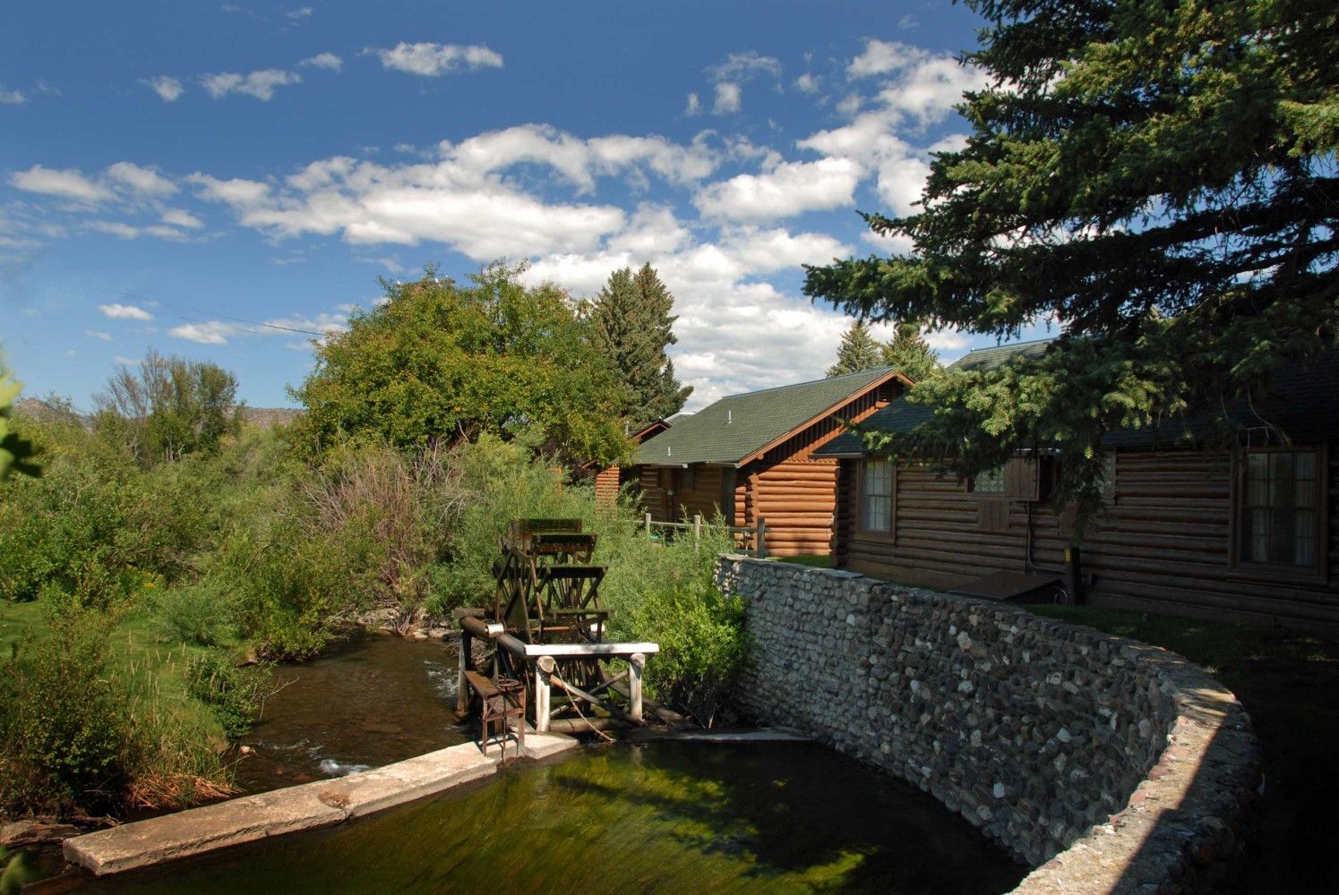 Garden pond beside a wooden cabin, with a stone wall, small waterwheel, and blue sky with clouds
