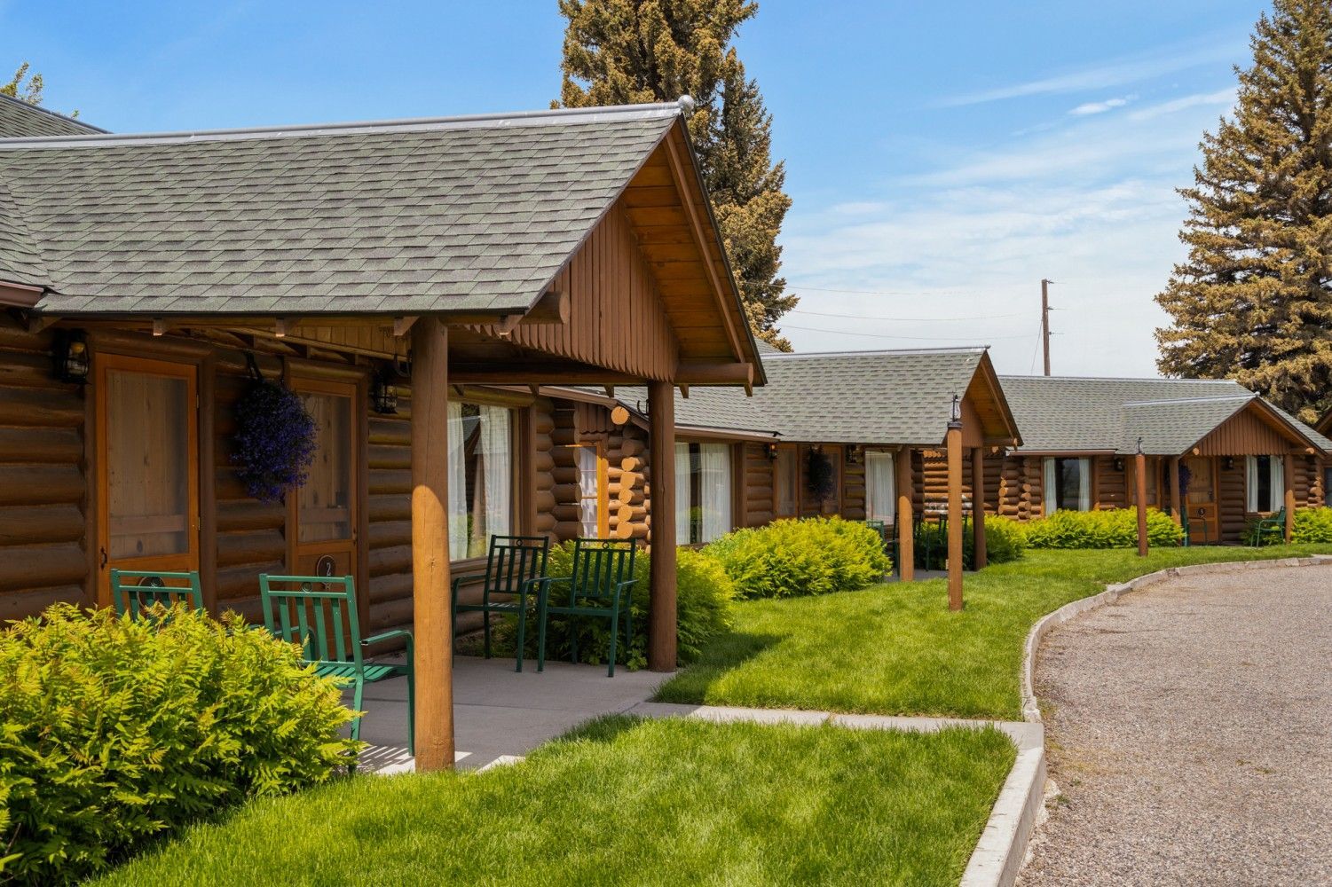 Log cabins along a gravel path with green lawns and shrubs under a clear blue sky