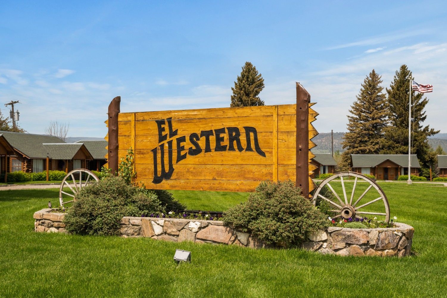 Wooden “The Western” sign on a grassy roadside with stone base and wagon wheel decor