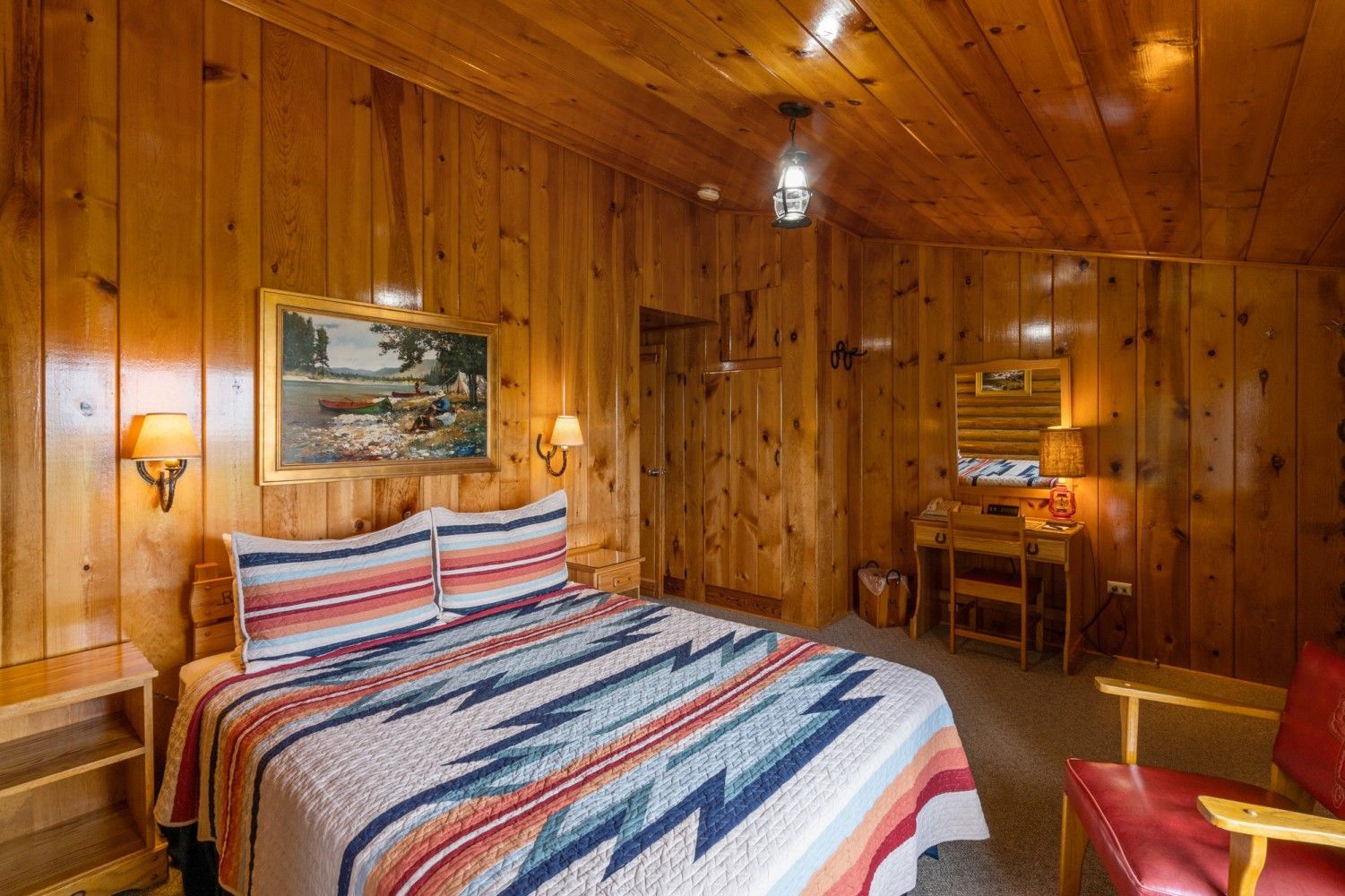 Cozy wood-paneled bedroom with a striped bedspread, wall art, lamp, and small desk