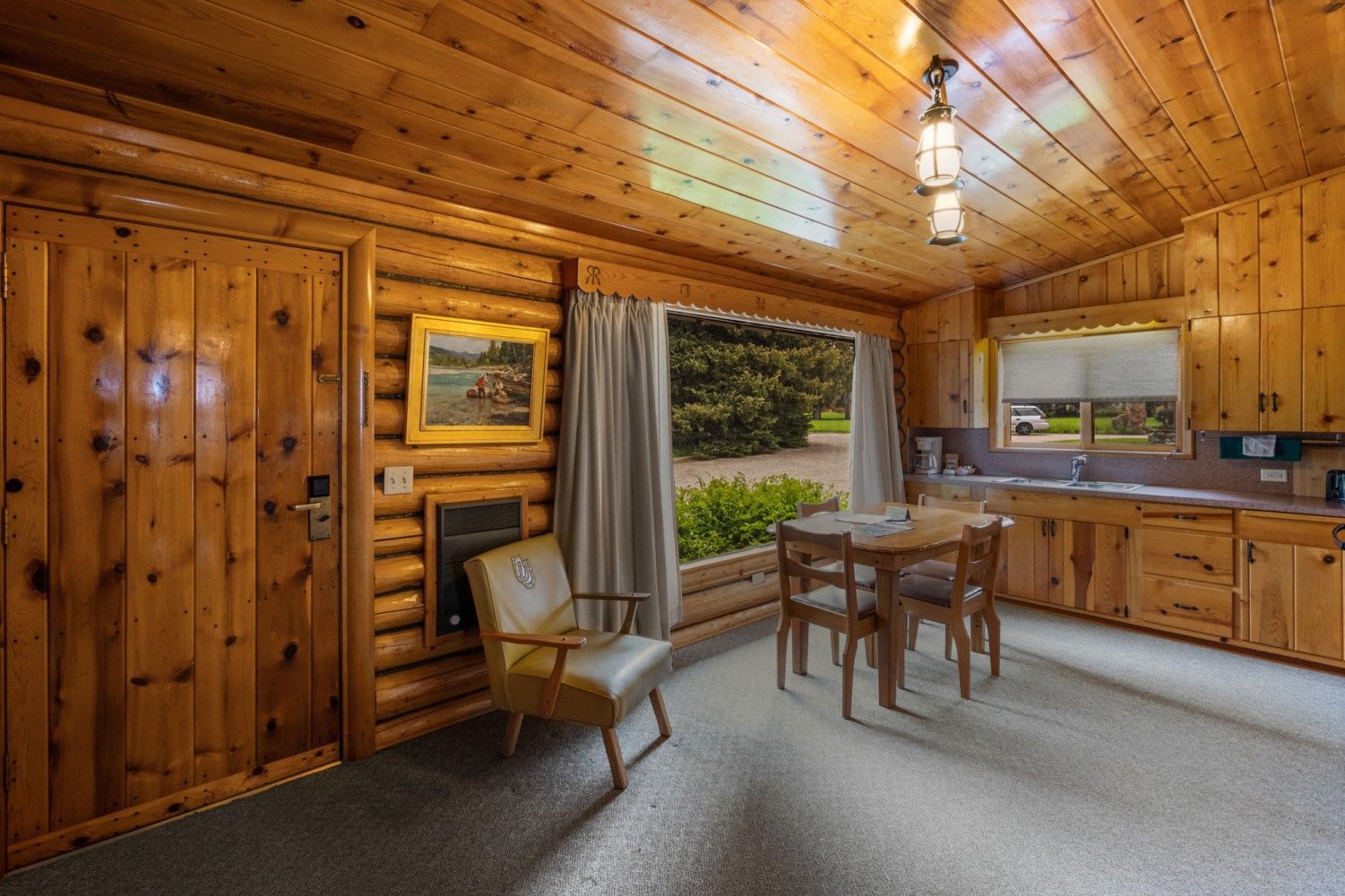 Cozy wood-paneled cabin interior with dining table, chairs, kitchenette, and large window view