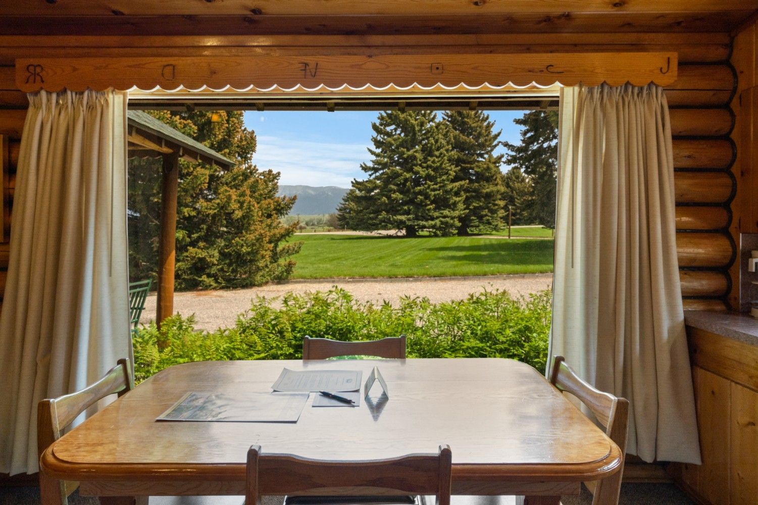 Dining table by open window with curtains, overlooking a green lawn and trees.