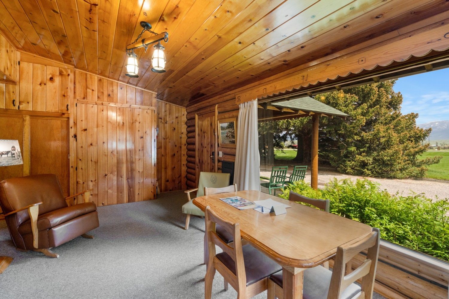 Wood-paneled cabin dining nook with table, leather chair, and large window overlooking greenery.