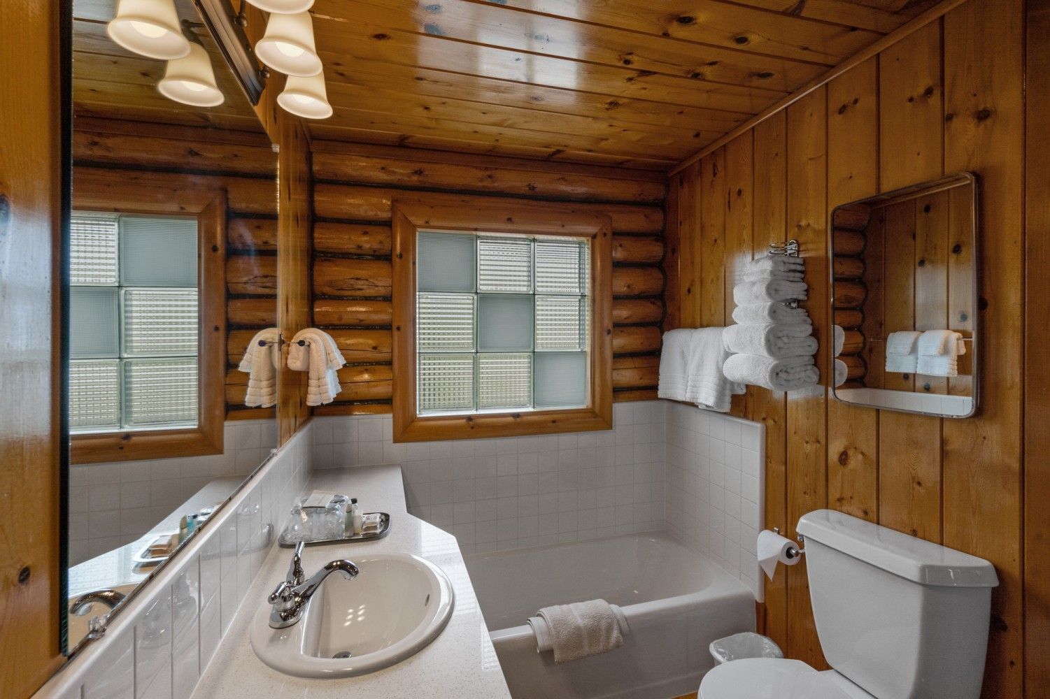Rustic wood-paneled bathroom with sink, toilet, shower-tub, and two windows