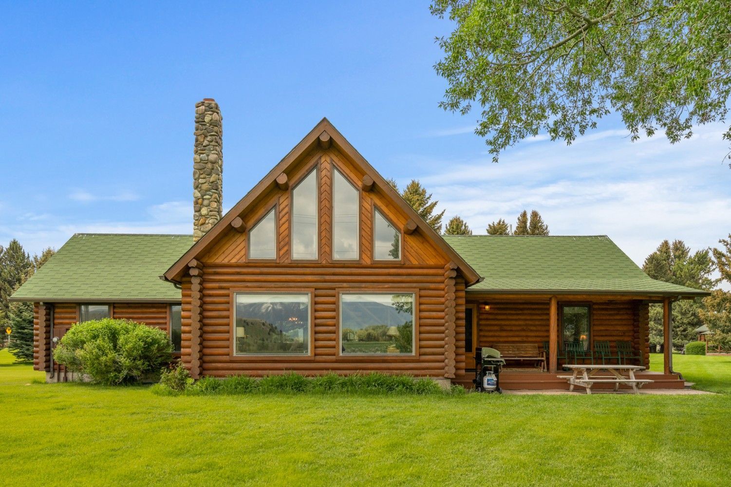Log cabin home with green roof, stone chimney, and large front windows in a grassy yard