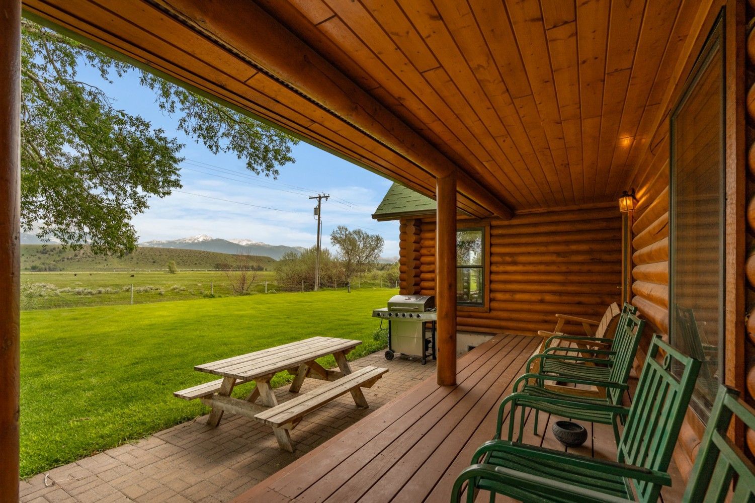 Covered log cabin porch with green chairs, picnic table, and grassy rural view beyond.