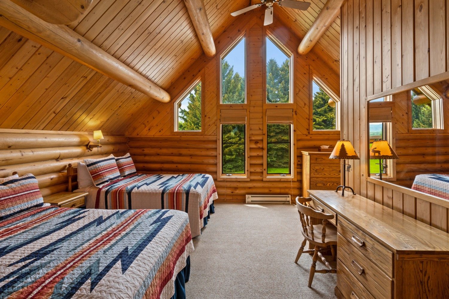 Cozy log cabin bedroom with two beds, desk, and large windows under a vaulted wooden ceiling