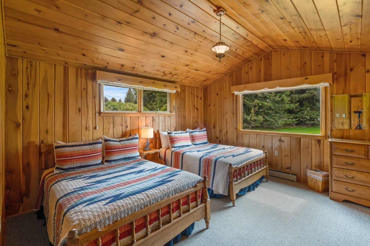 Wood-paneled bedroom with two twin beds, plaid bedding, and large windows overlooking trees