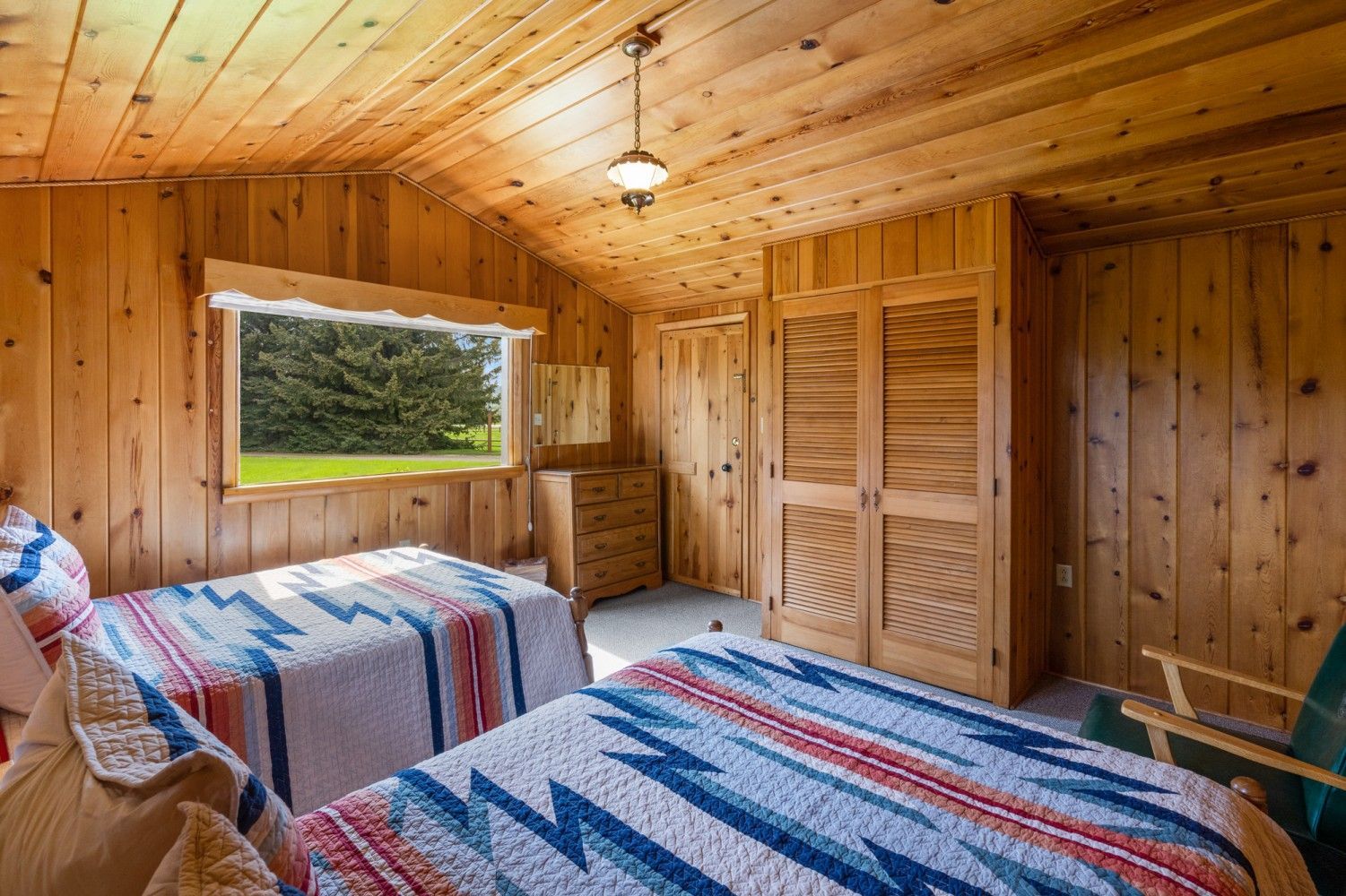 Cozy wood-paneled bedroom with two twin beds, plaid quilts, and a window overlooking trees