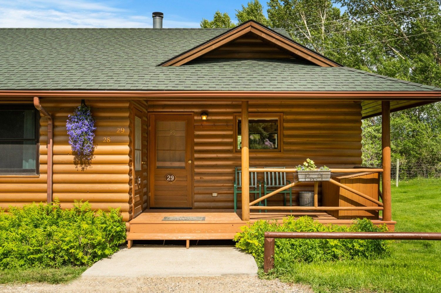 Log cabin with a wooden porch, green roof, and hanging purple flowers surrounded by grass.