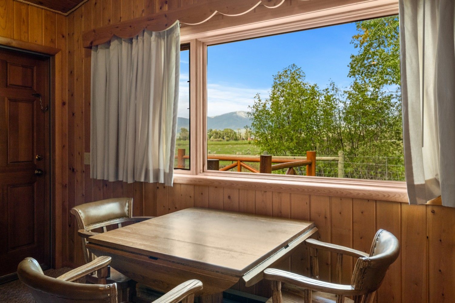 Wood-paneled dining nook with a table and chairs overlooking a sunny mountain landscape through a large window