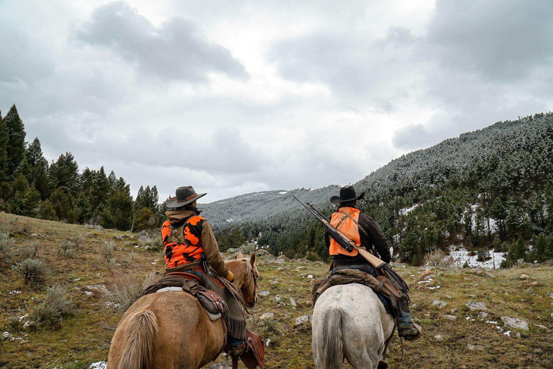 Two riders in orange vests horseback ride through a rocky mountain meadow under cloudy skies