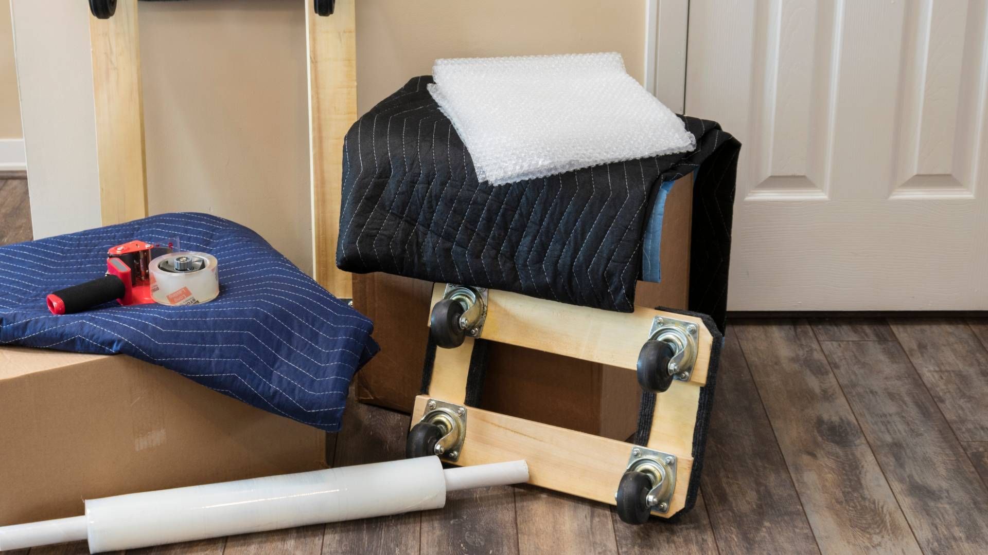 Moving supplies: box on a dolly, packing blankets, tape, plastic wrap, and bubble wrap, near a doorway.