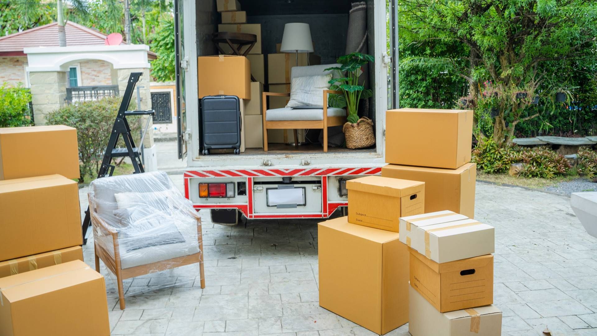 Moving truck open, filled with boxes and furniture. Boxes and a chair outside. Backyard setting.