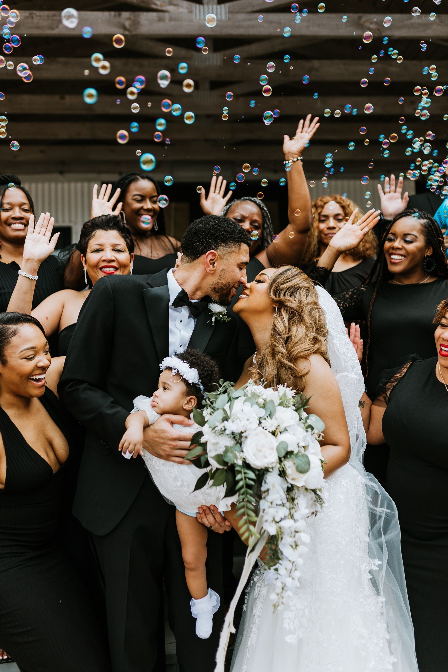 A bride and groom are kissing while their wedding party throws soap bubbles in the air.