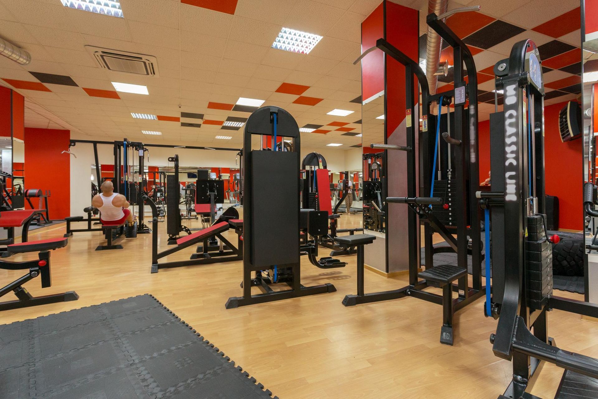 Gym interior with weight machines, mirrors, and a person sitting by the wall