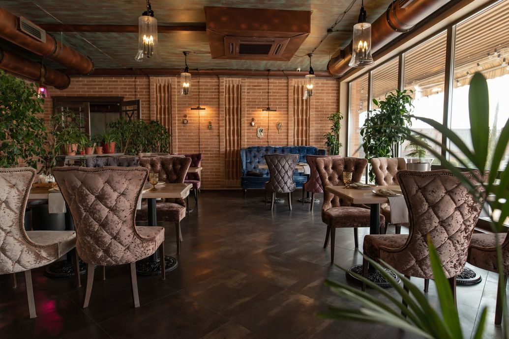 Cozy restaurant interior with tufted chairs, wooden tables, warm lighting, and plants by large windows