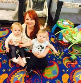 A smiling red-haired person sits on a colorful, patterned rug with two babies, one on each lap, in a play area.