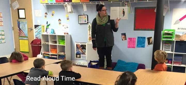 A teacher stands in a classroom reading a book aloud to a group of young children sitting at desks.