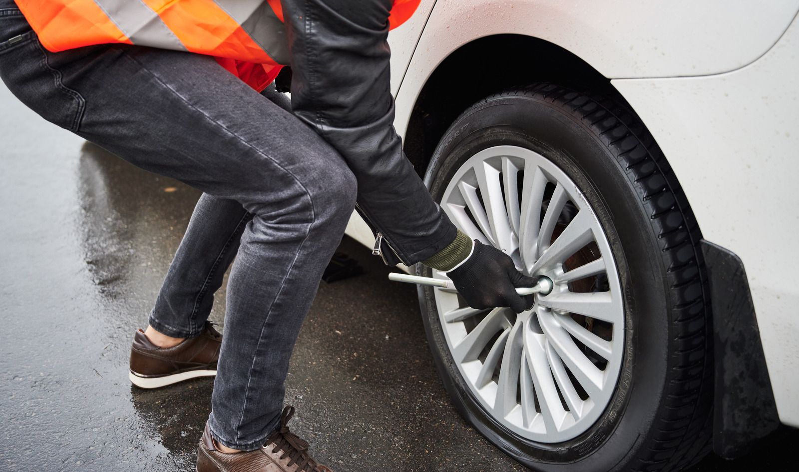 Worker using a wrench to remove a car wheel in the rain, wearing a reflective safety vest.