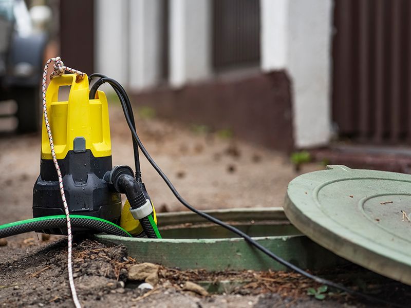 Yellow and black submersible pump sitting next to an open green access hatch. A green hose and black power cord are attached. Yellow and black submersible pump sitting next to an open green access hatch. A green hose and black power cord are attached.