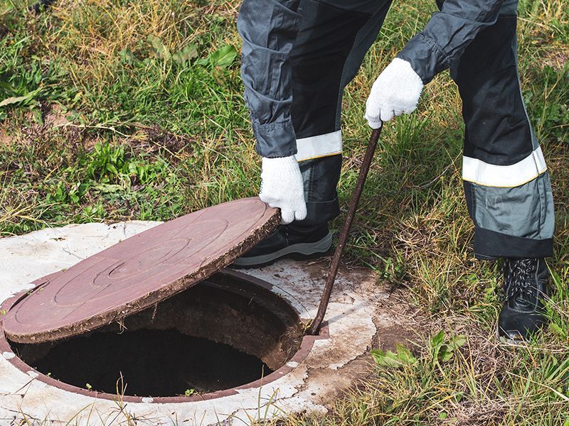 Person in work clothes lifting a manhole cover with a hook in a grassy area. Person in work clothes lifting a manhole cover with a hook in a grassy area.