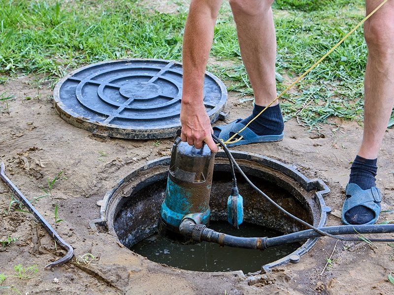 Person removing a pump from a septic tank. The open tank is in a dirt yard, the lid nearby. Person removing a pump from a septic tank. The open tank is in a dirt yard, the lid nearby.