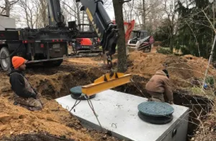 Workers installing a septic tank, with a crane lowering it into a dug pit. Dirt and construction vehicles surround them, in an outdoor setting.