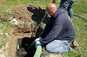 A man kneels beside an open septic tank, holding a large hose. He appears to be working on or pumping the tank.