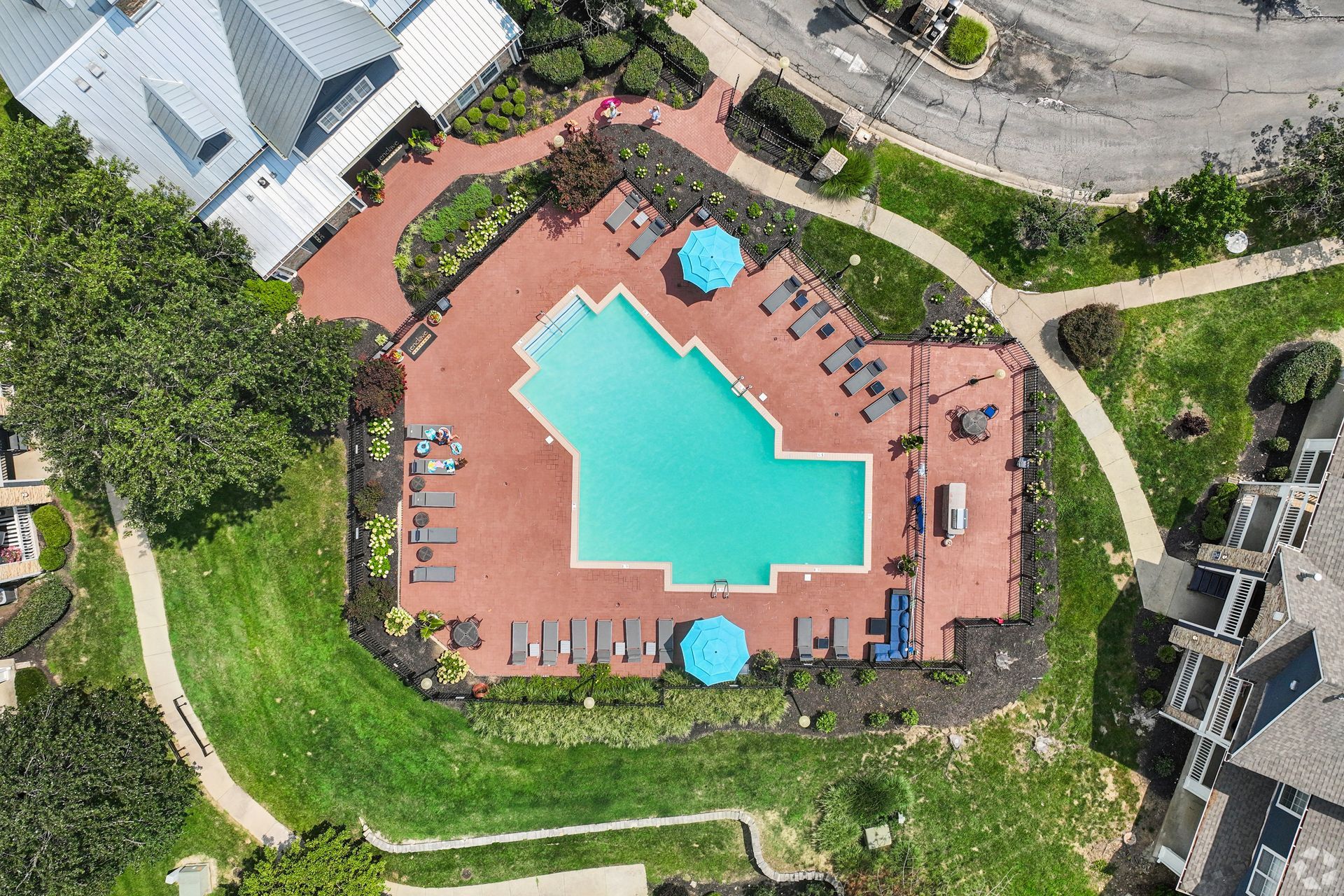 Aerial view of a turquoise swimming pool surrounded by a brick patio, lounge chairs, umbrellas, and greenery.