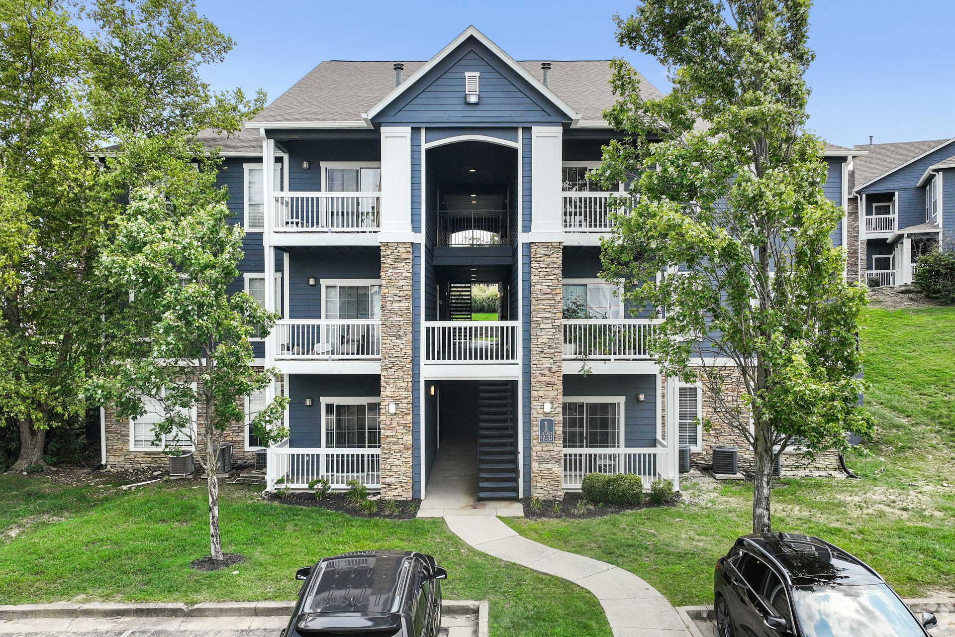 Three-story apartment building with balconies, trees, and parked cars in front