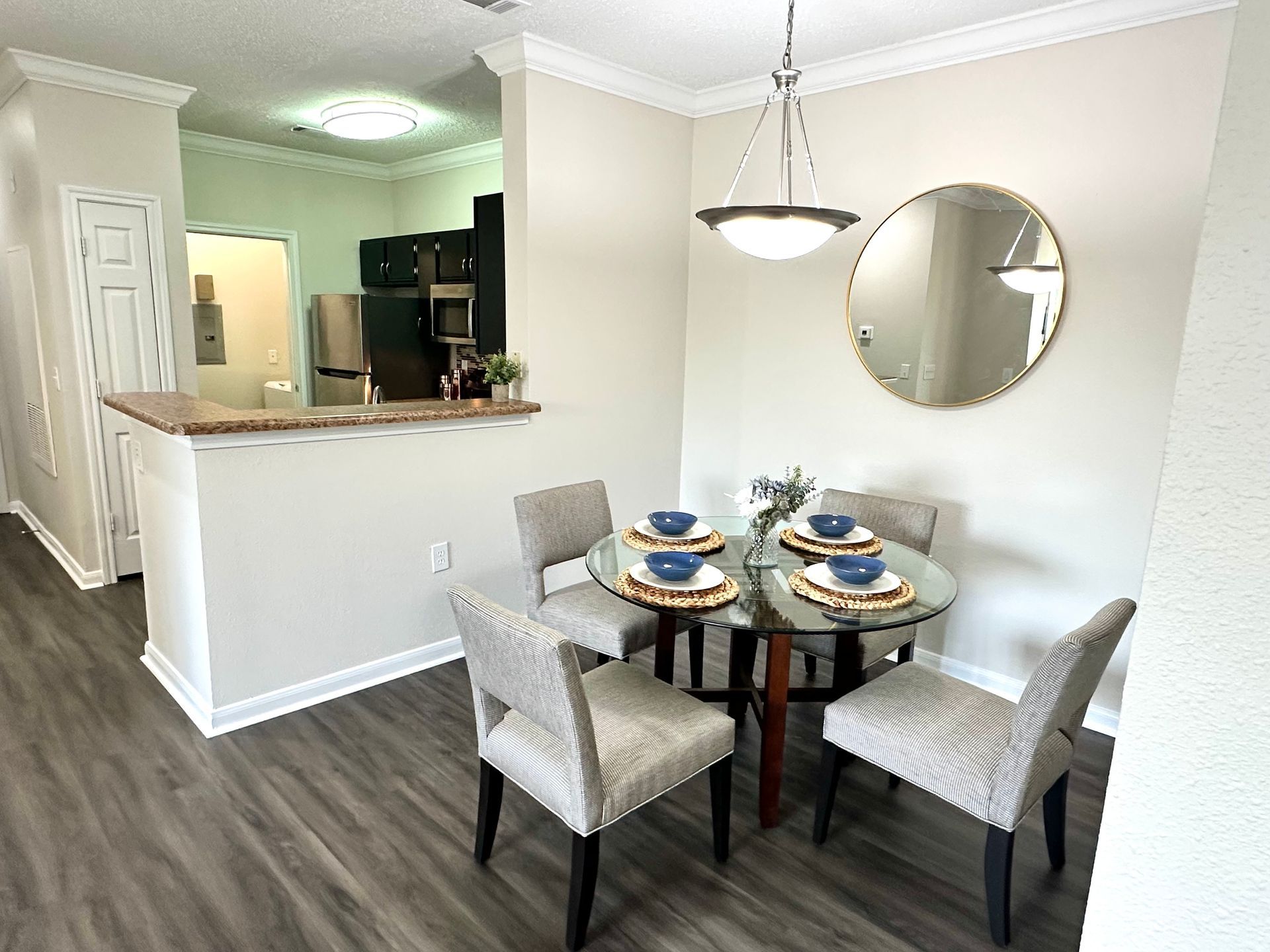 Dining area with round glass table, four chairs, and a mirror. Light gray walls, dark wood floor.
