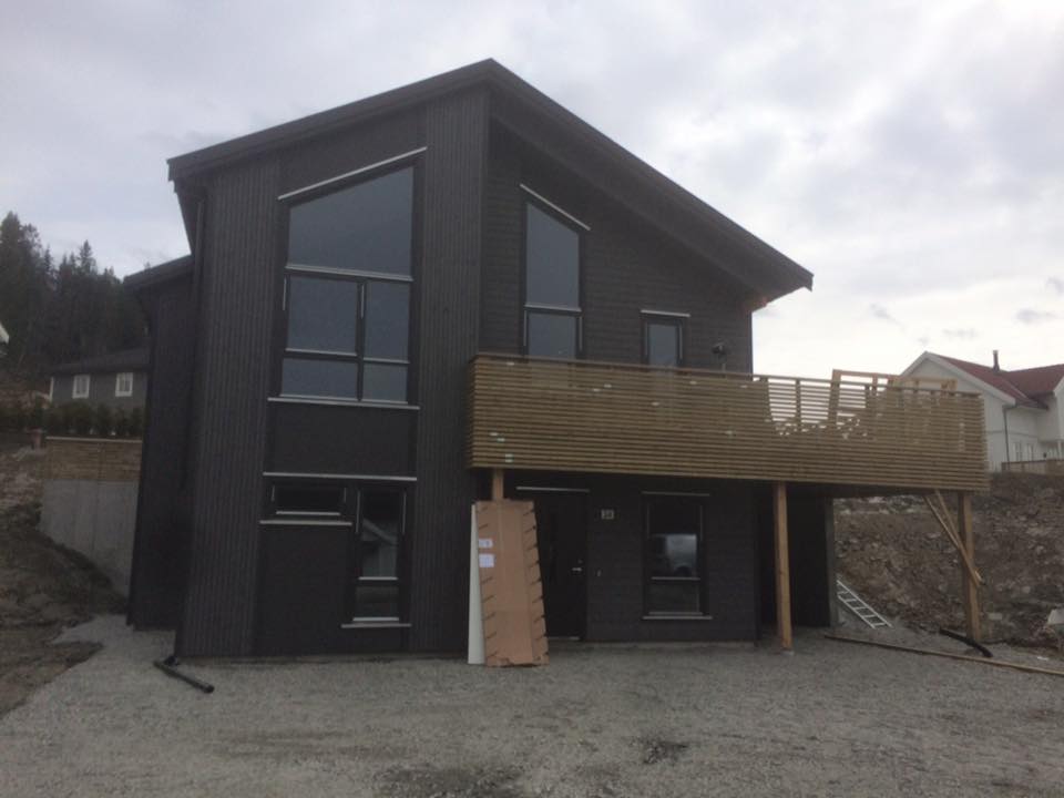 Moderne mørkegrått hus med store vinduer, trebalkong og steinstøttemurer under en overskyet himmel