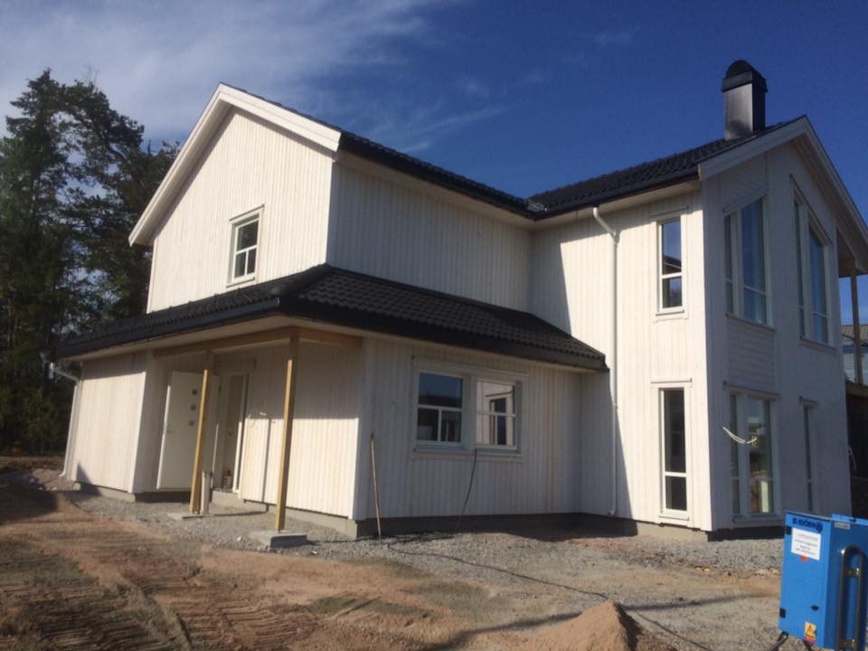 Toetasjes hvitt hus med mørkt tak og veranda, vist i dagslys på en grustomt