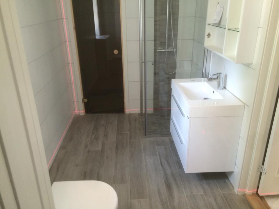 Smalt moderne bad med grå fliser, hvitt servantskap, glassdusj og toalett i forgrunnen