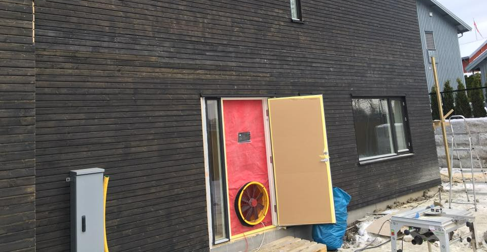 Husets eksteriør med mørk ytterkledning, rød dør og byggematerialer ved siden av