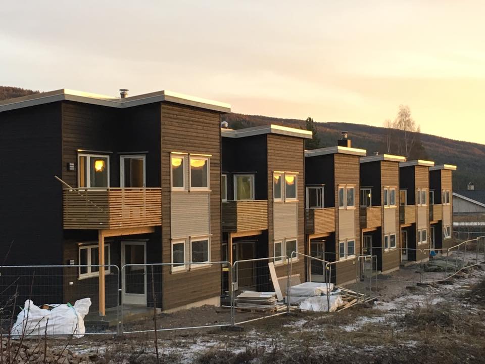 Rekke med moderne brune leilighetsenheter under bygging ved solnedgang, med snødekt terreng og fjell bak.